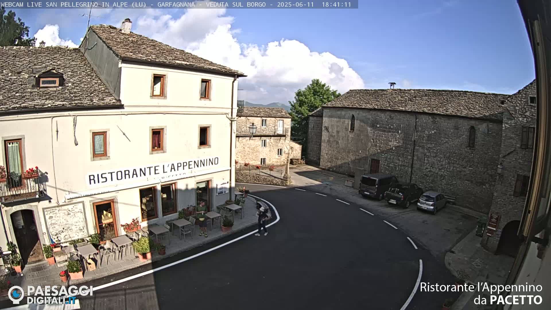A partly cloudy day reveals a quaint town square with stone buildings, a restaurant with outdoor seating, and a few parked cars.