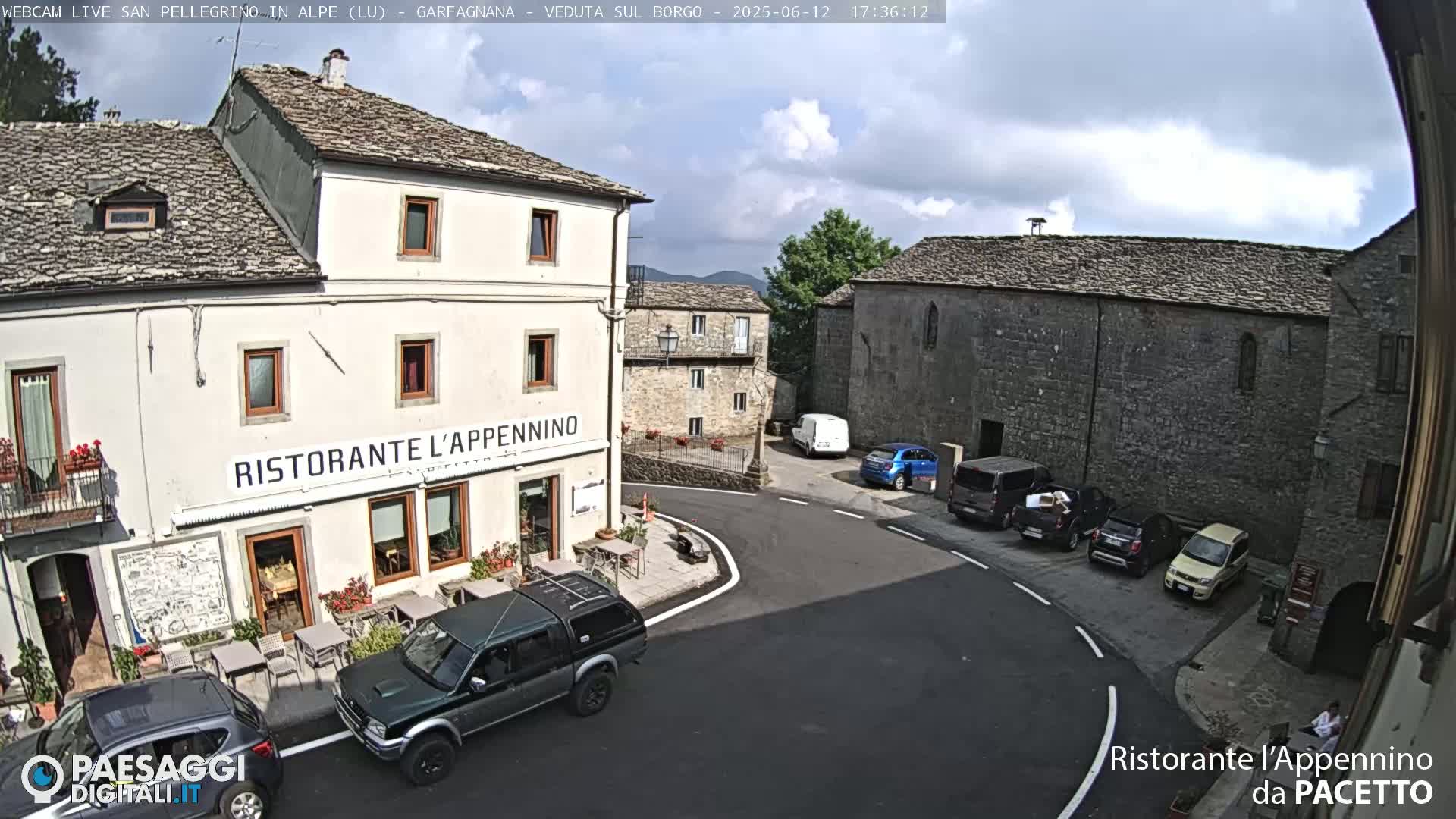A partly cloudy day in a stone village square shows several parked cars near a restaurant with outdoor seating.