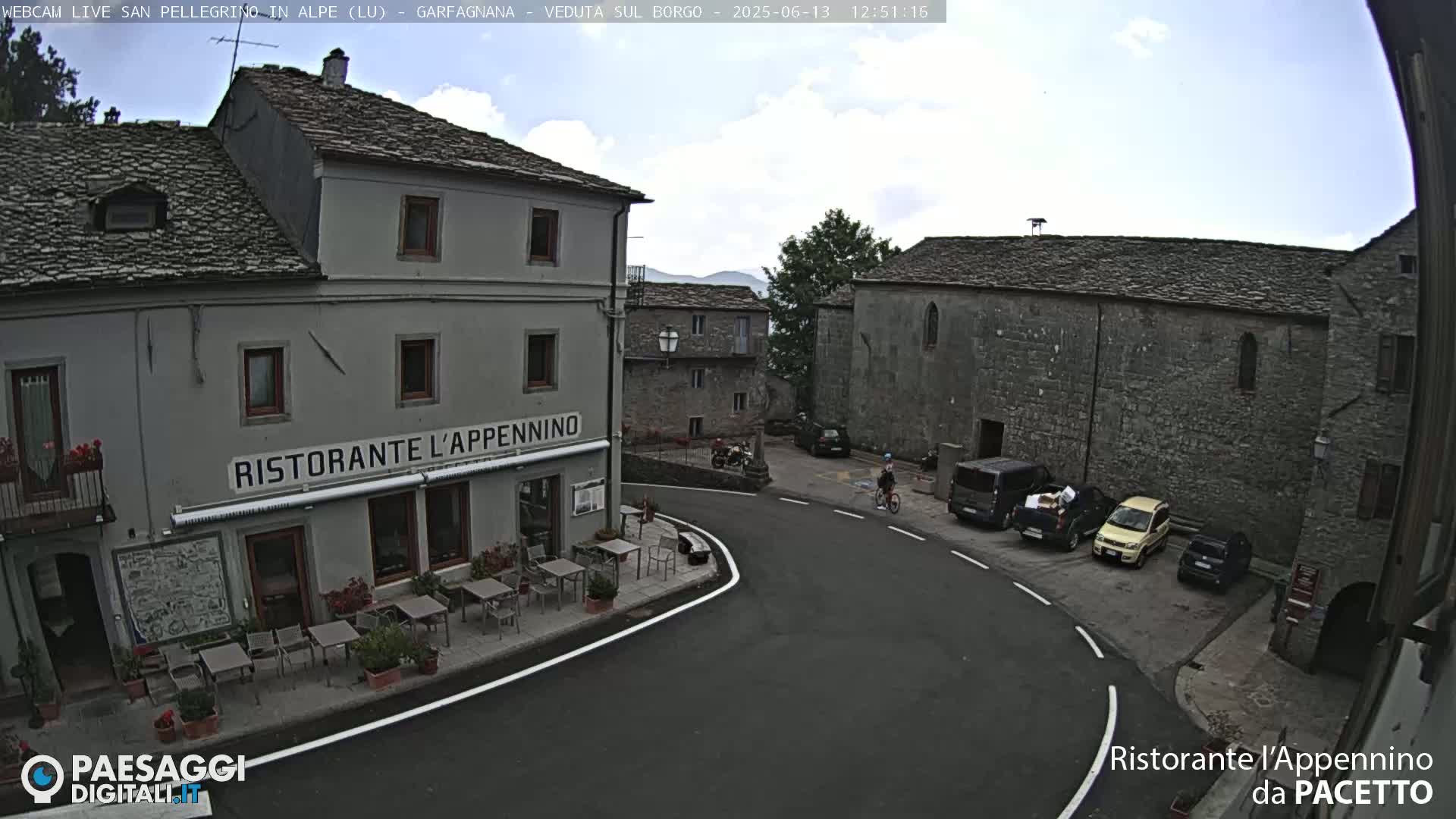 A partly cloudy day reveals a stone village square with a restaurant, several parked cars, and a person riding a bicycle.
