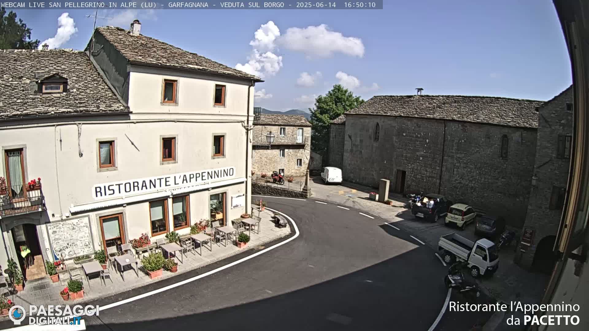 A sunny day in a quaint Italian village shows a restaurant with outdoor seating situated at a street corner next to a stone building, with several parked cars visible.