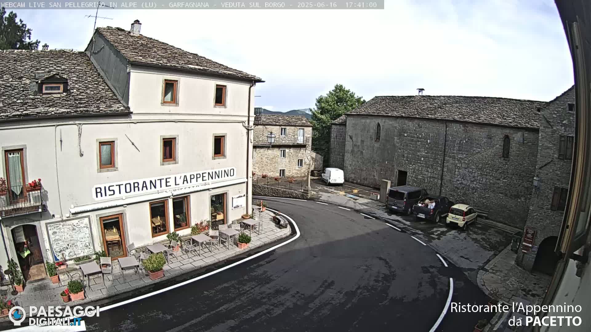 A high-angle view shows a quaint village square on a partly sunny day, featuring stone buildings, a restaurant with outdoor seating, and several parked cars.
