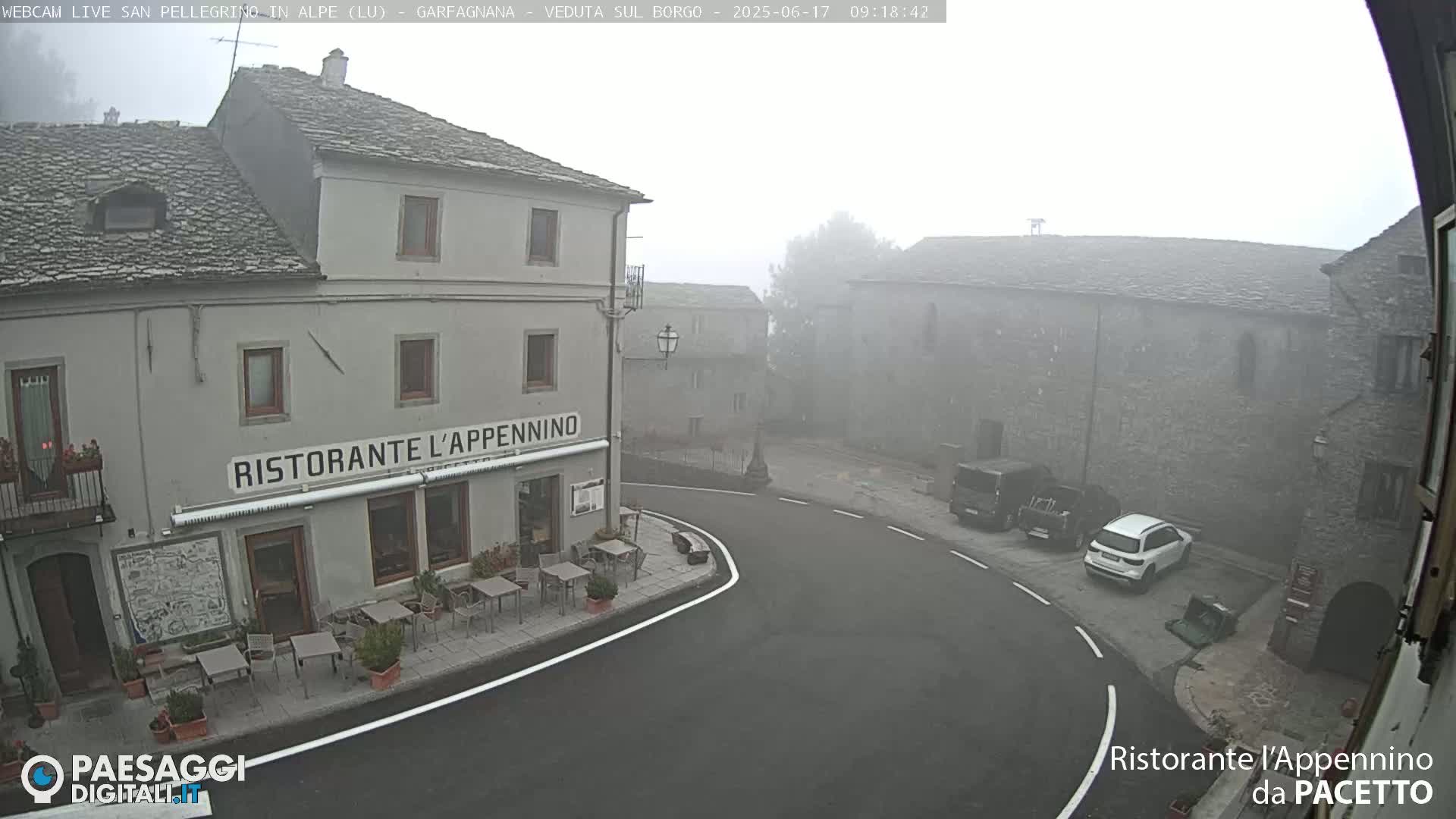 A foggy village square features a restaurant with outdoor seating and several parked vehicles.