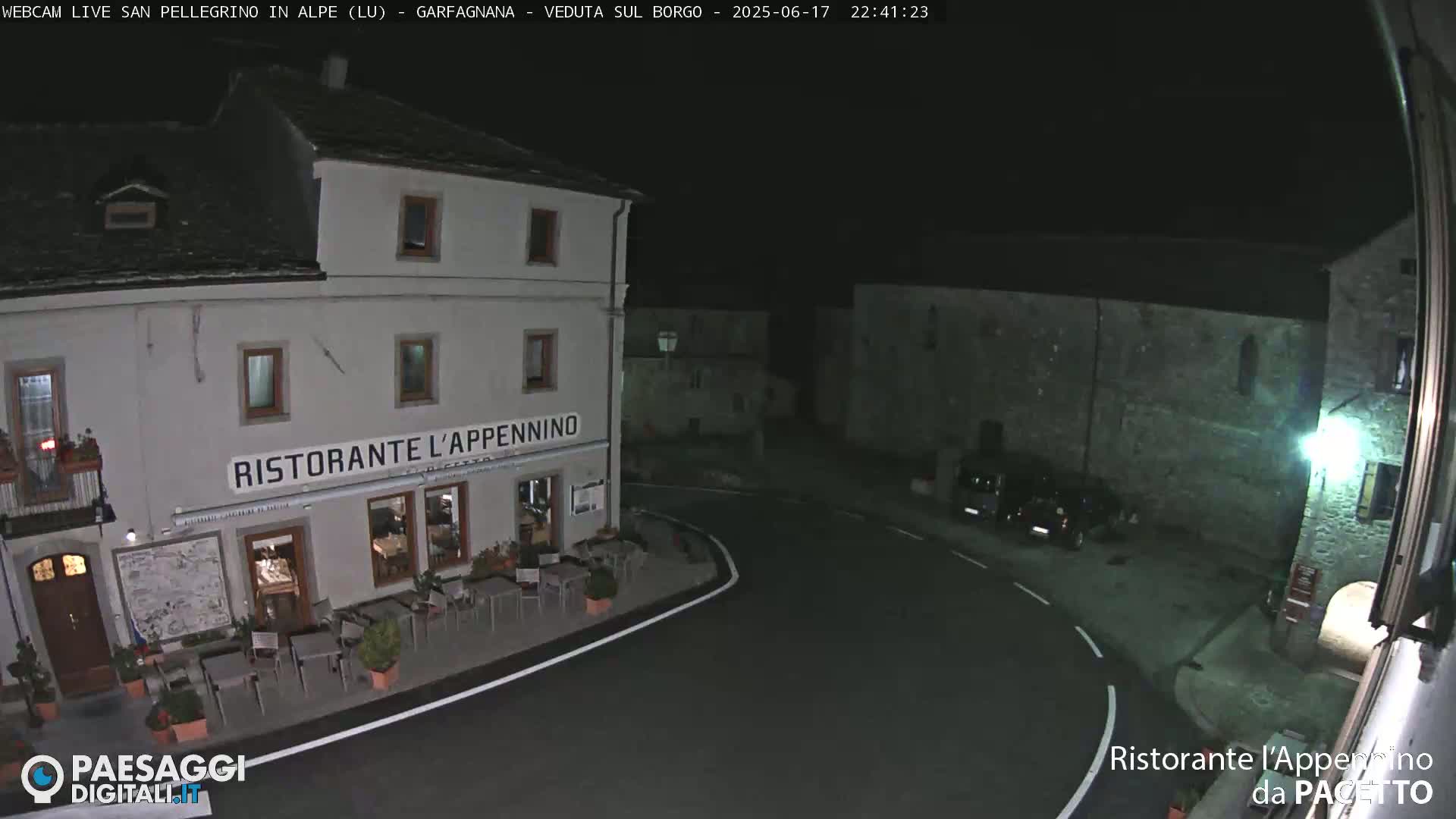 A nighttime, high-angle view of a town square with a restaurant, several parked cars, and stone buildings under a clear, dark sky.