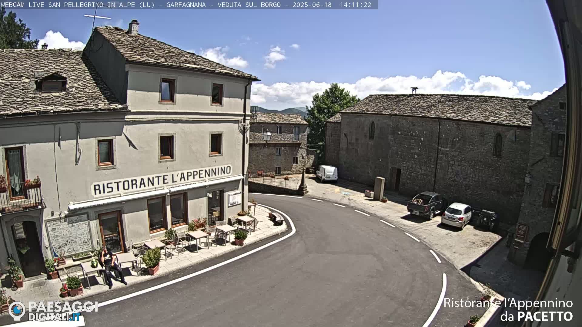 A sunny day reveals a quaint town square with stone buildings, a restaurant with outdoor seating, and a few parked cars.