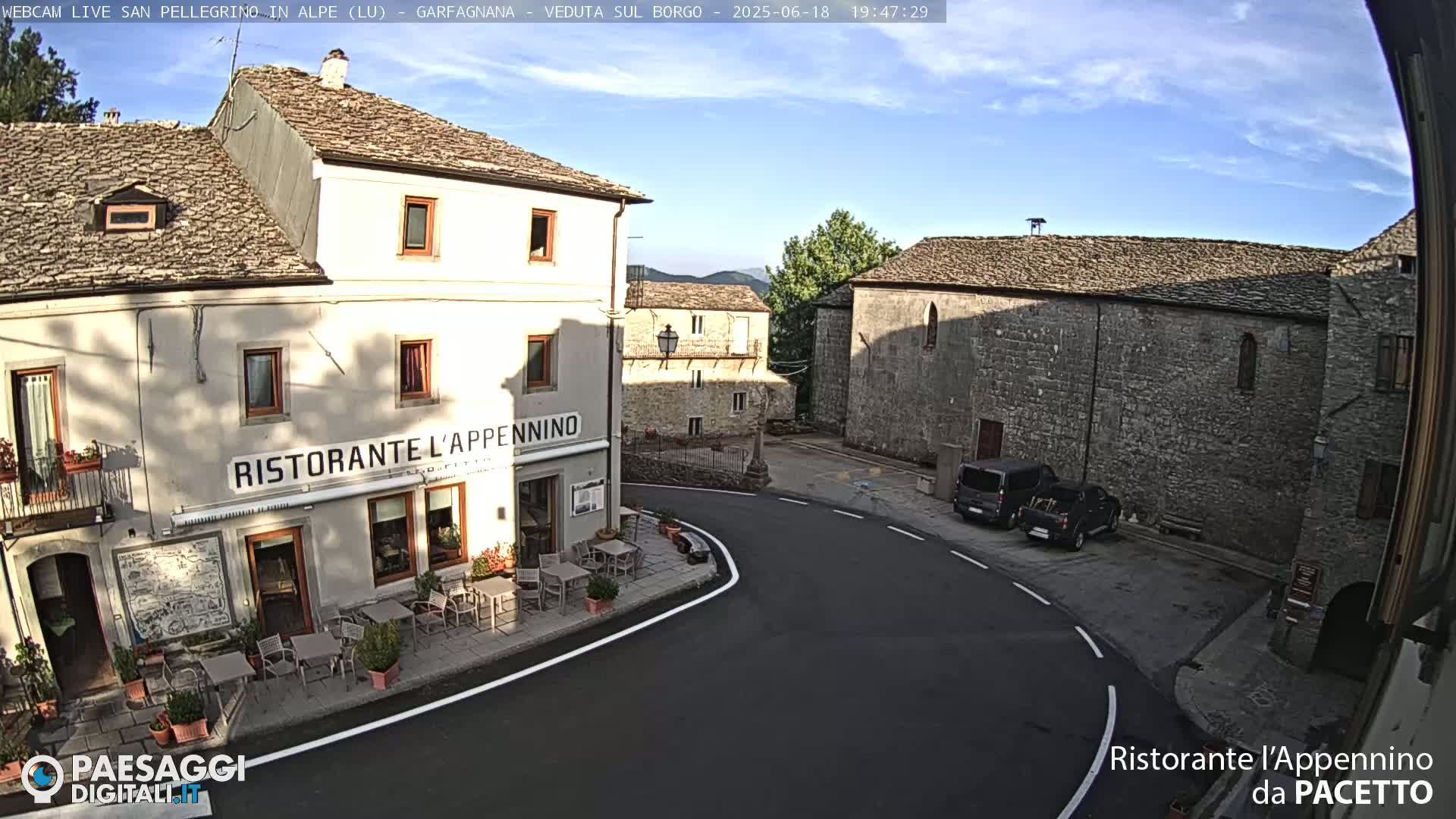 A restaurant with outdoor seating is situated on a corner in a stone village under a mostly clear sky.