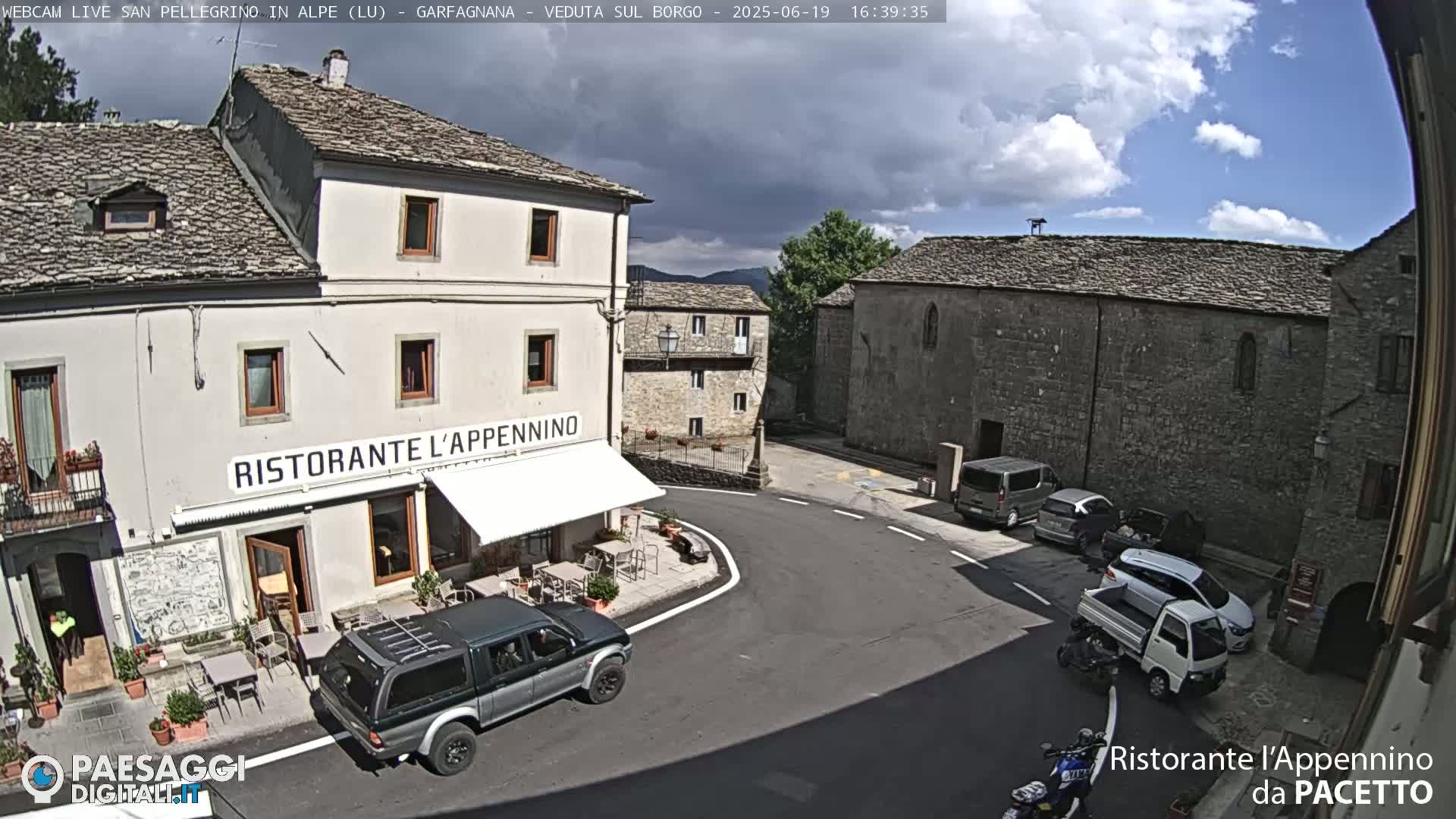 A partly cloudy daytime view of a town square with several parked vehicles, a restaurant with outdoor seating, and stone buildings.