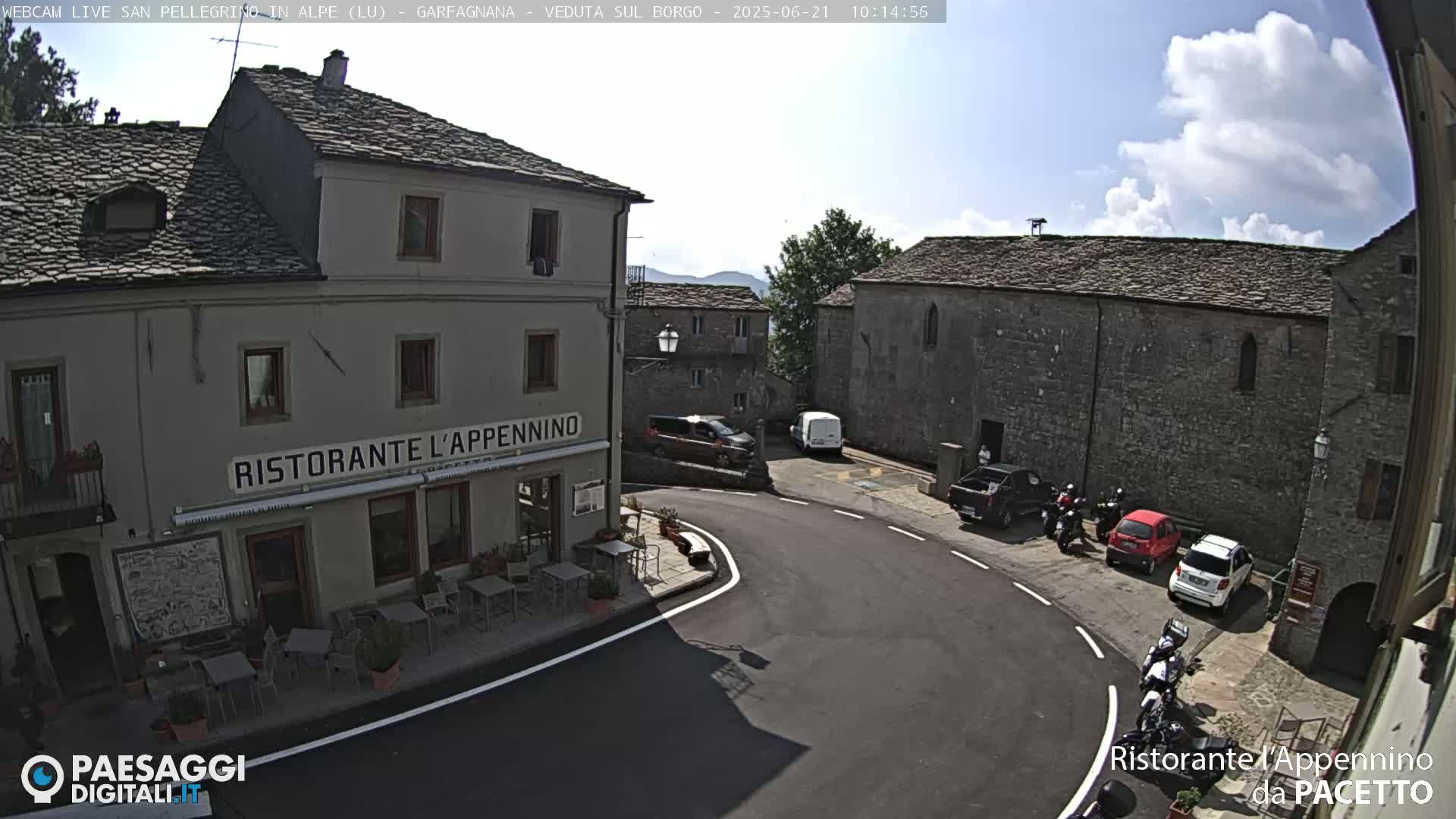 A partly sunny day reveals a quaint village square with stone buildings, parked cars and motorcycles, and a restaurant with outdoor seating.