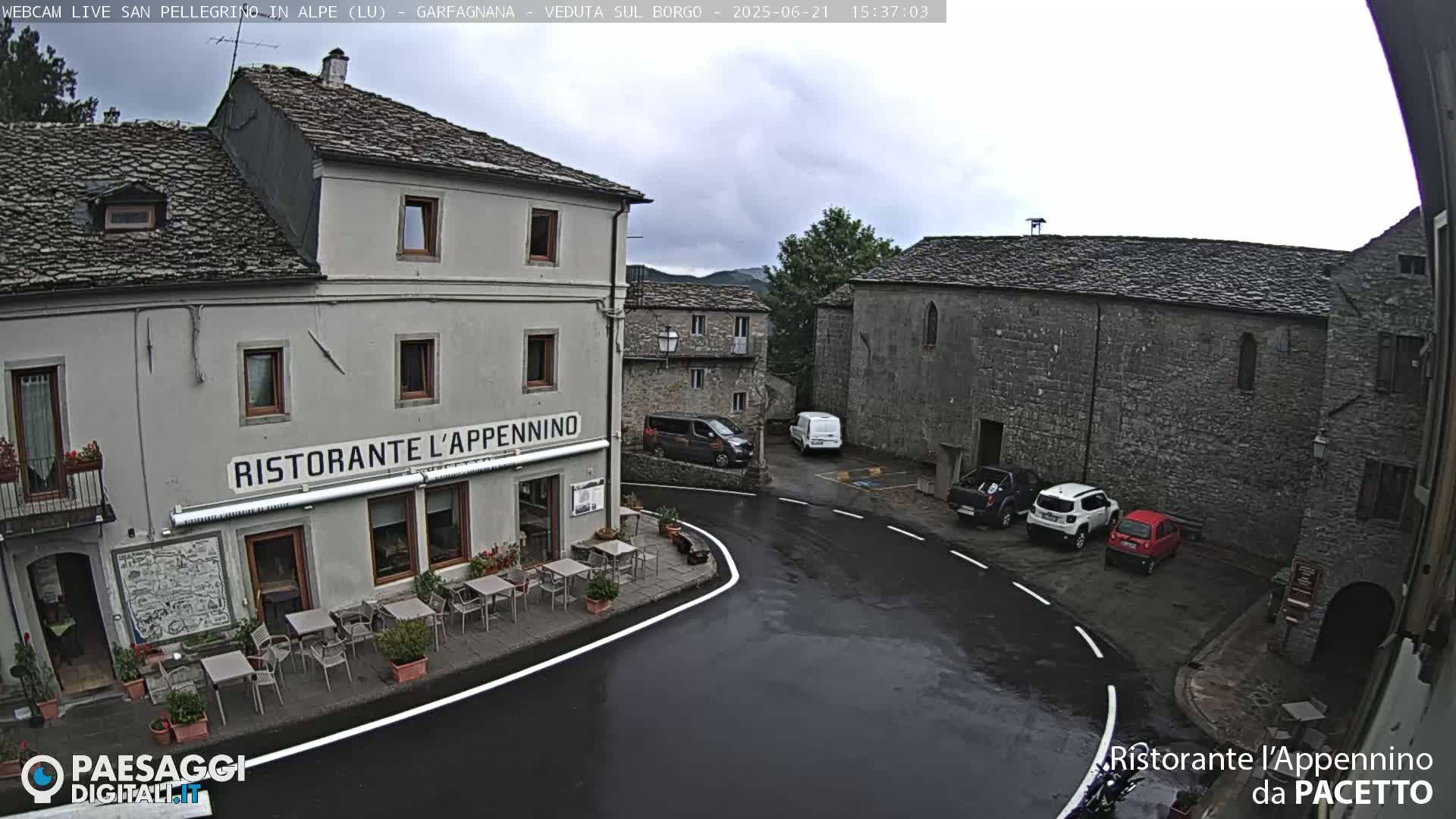 A wet village square with a restaurant, several parked cars, and stone buildings under an overcast sky.