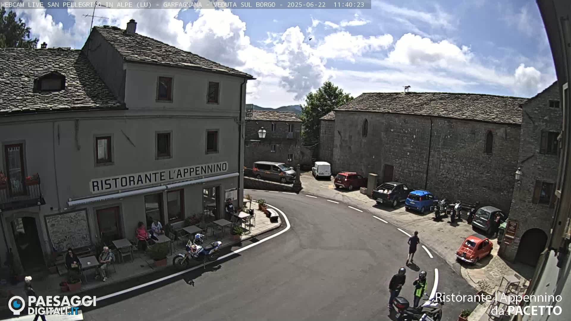 A sunny village square with stone buildings, parked cars and motorcycles, and people sitting at outdoor tables in front of a restaurant.