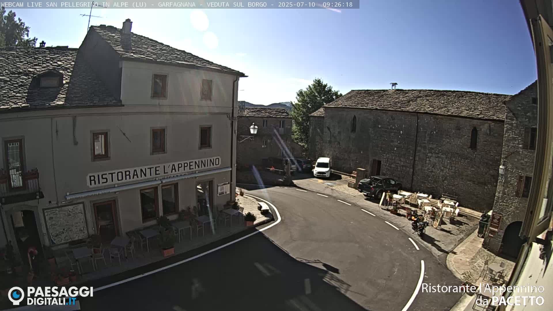A sunny outdoor view of a town square with stone buildings, a restaurant with outdoor seating, and several parked vehicles.