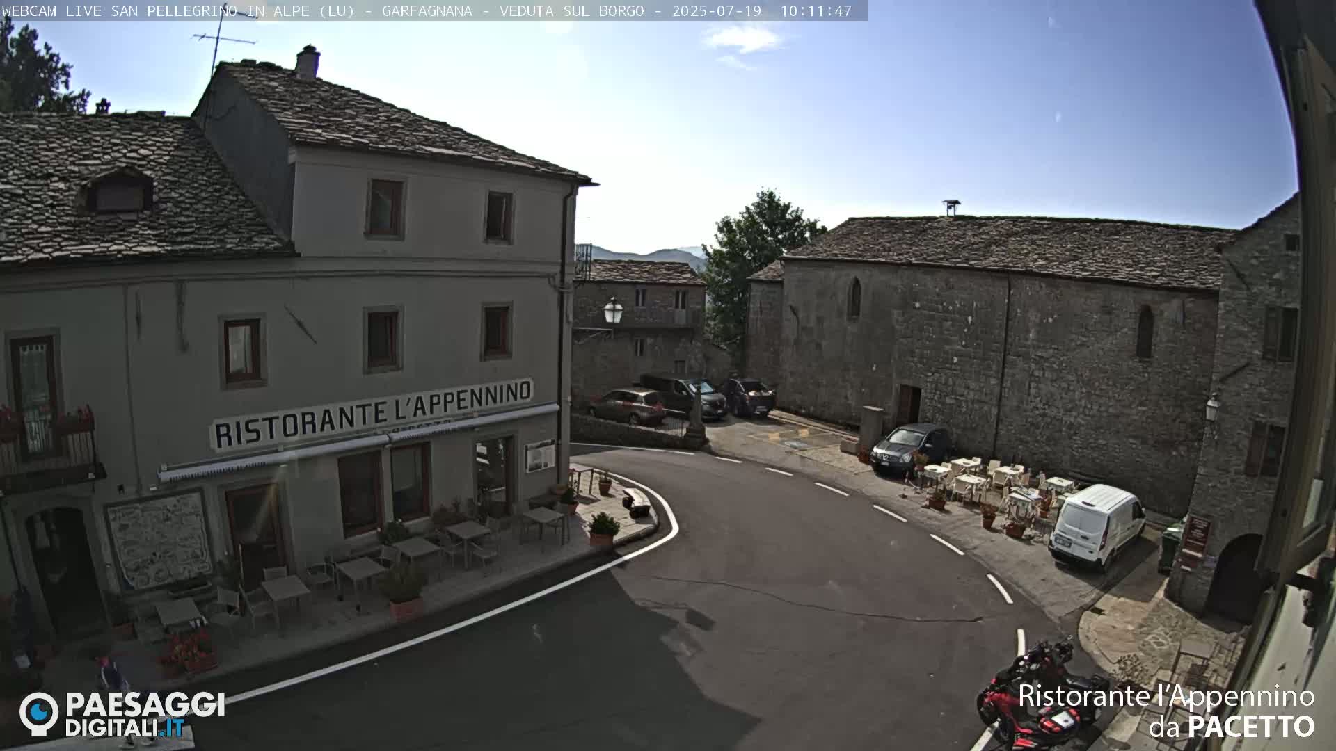 A sunny view of a town square with several parked cars, a restaurant with outdoor seating, and stone buildings.