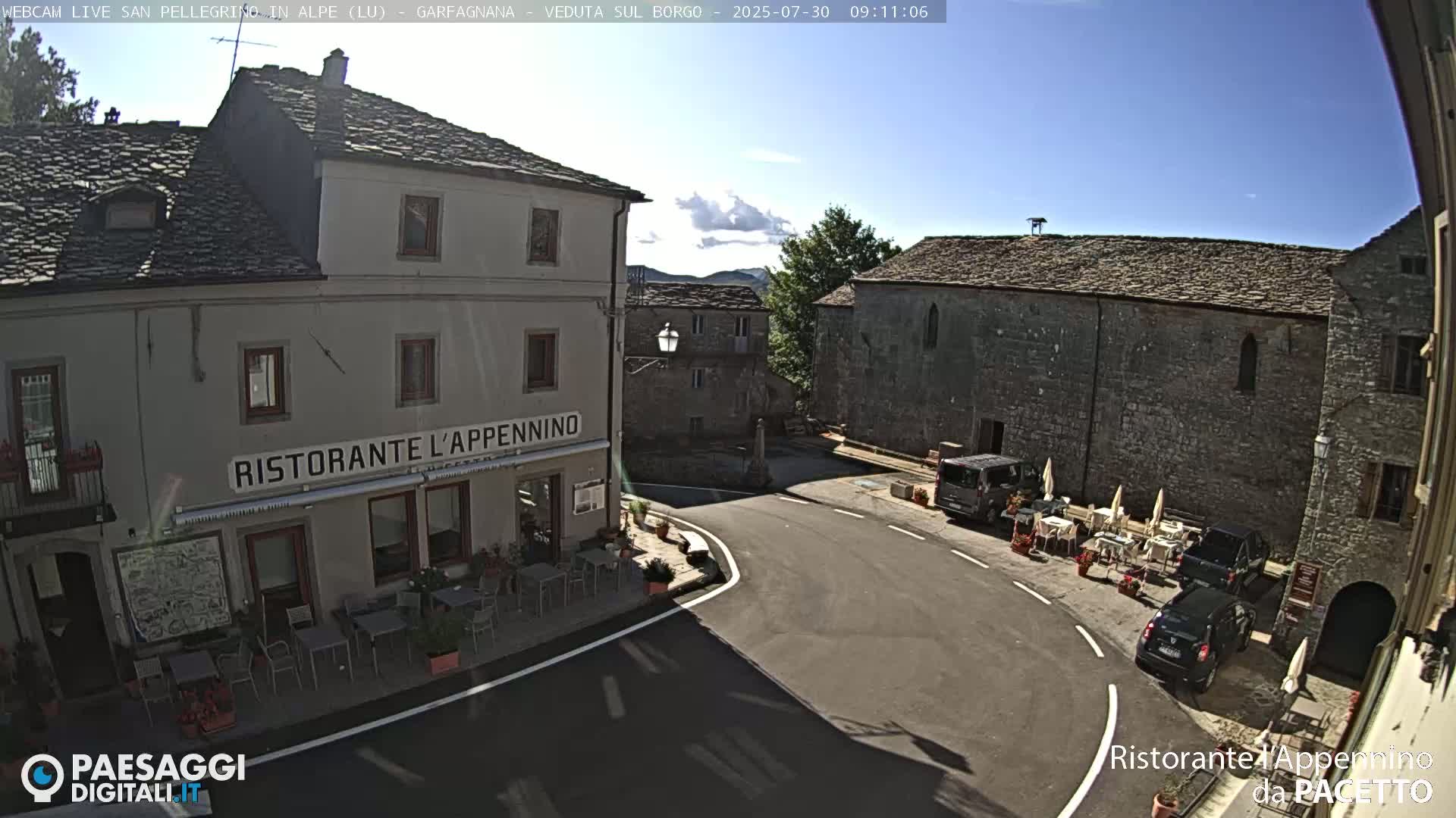 A sunny day reveals a town square with a restaurant, parked cars, and outdoor seating, surrounded by stone buildings.