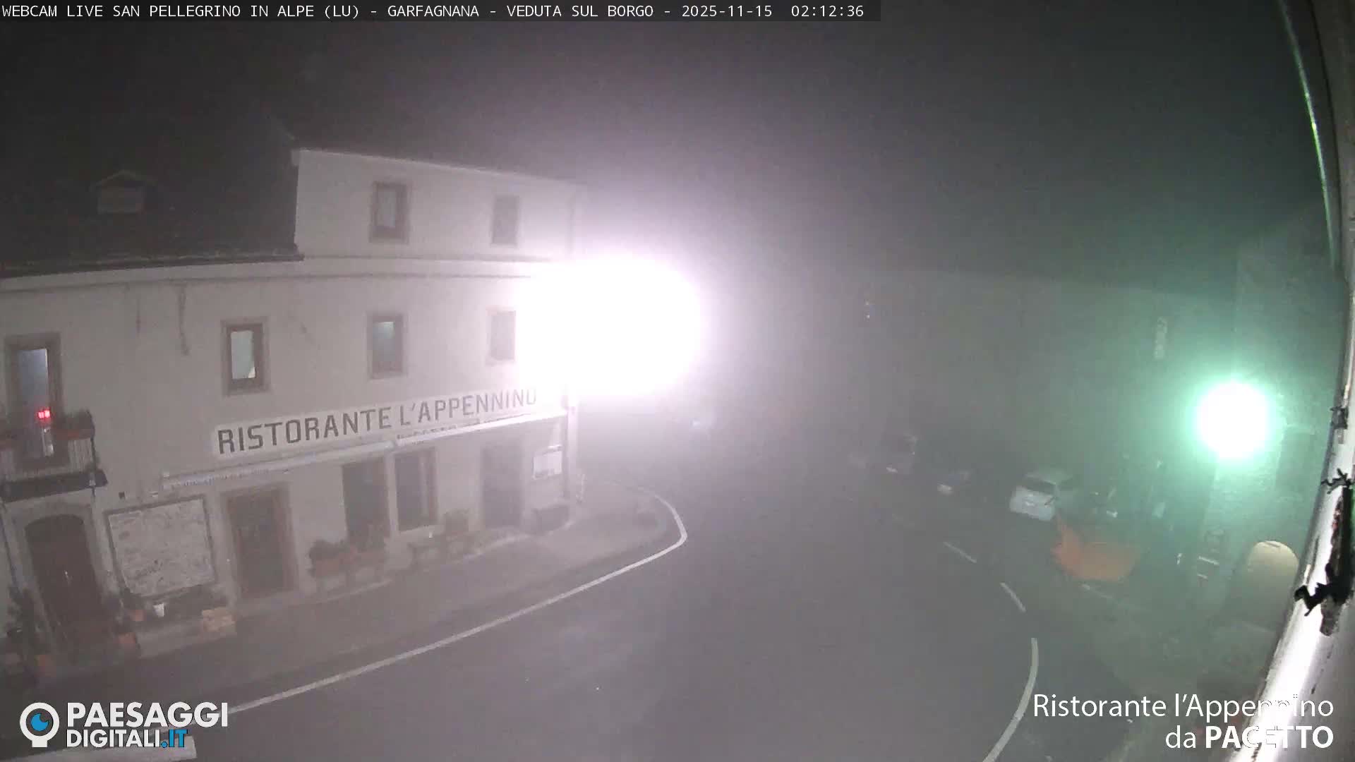 A village street is seen at night through dense fog, illuminated by intense streetlights that create a bright glare in front of a two-story restaurant building.
