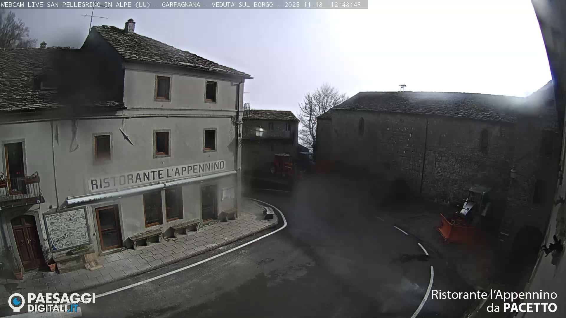 A village street is seen at night through dense fog, illuminated by intense streetlights that create a bright glare in front of a two-story restaurant building.