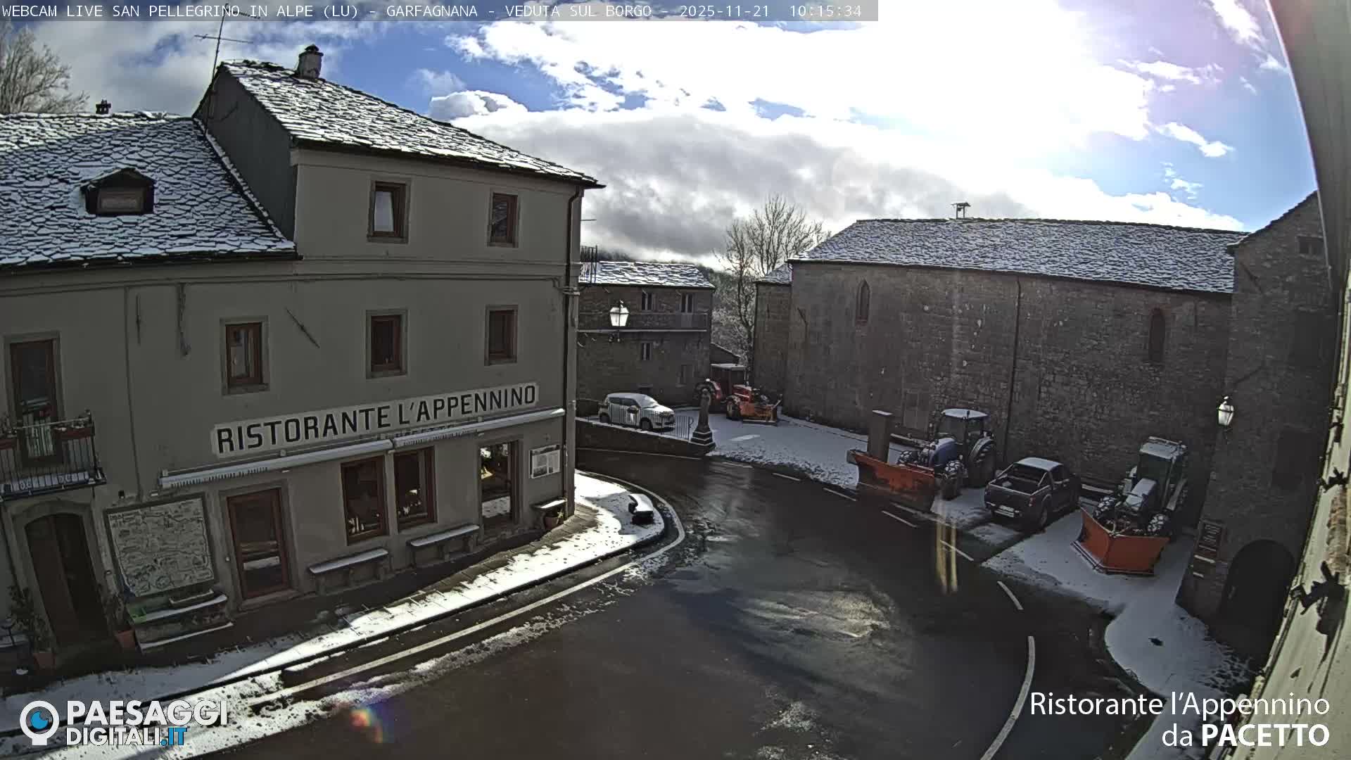 A wintry view of a historic European village square shows snow-dusted stone buildings, wet roads, and parked snowplows under a bright, partly cloudy sky.