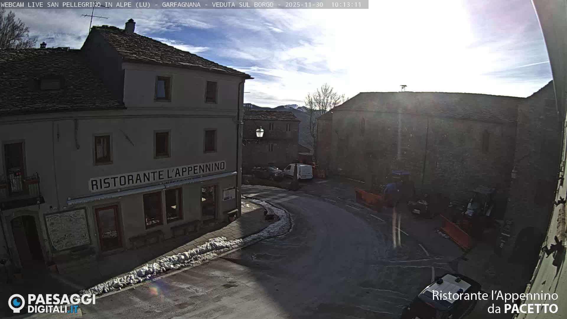 A quaint village street, partially covered in snow on the sidewalks, winds past stone buildings including a restaurant on a bright, partly cloudy day with distant mountains visible.