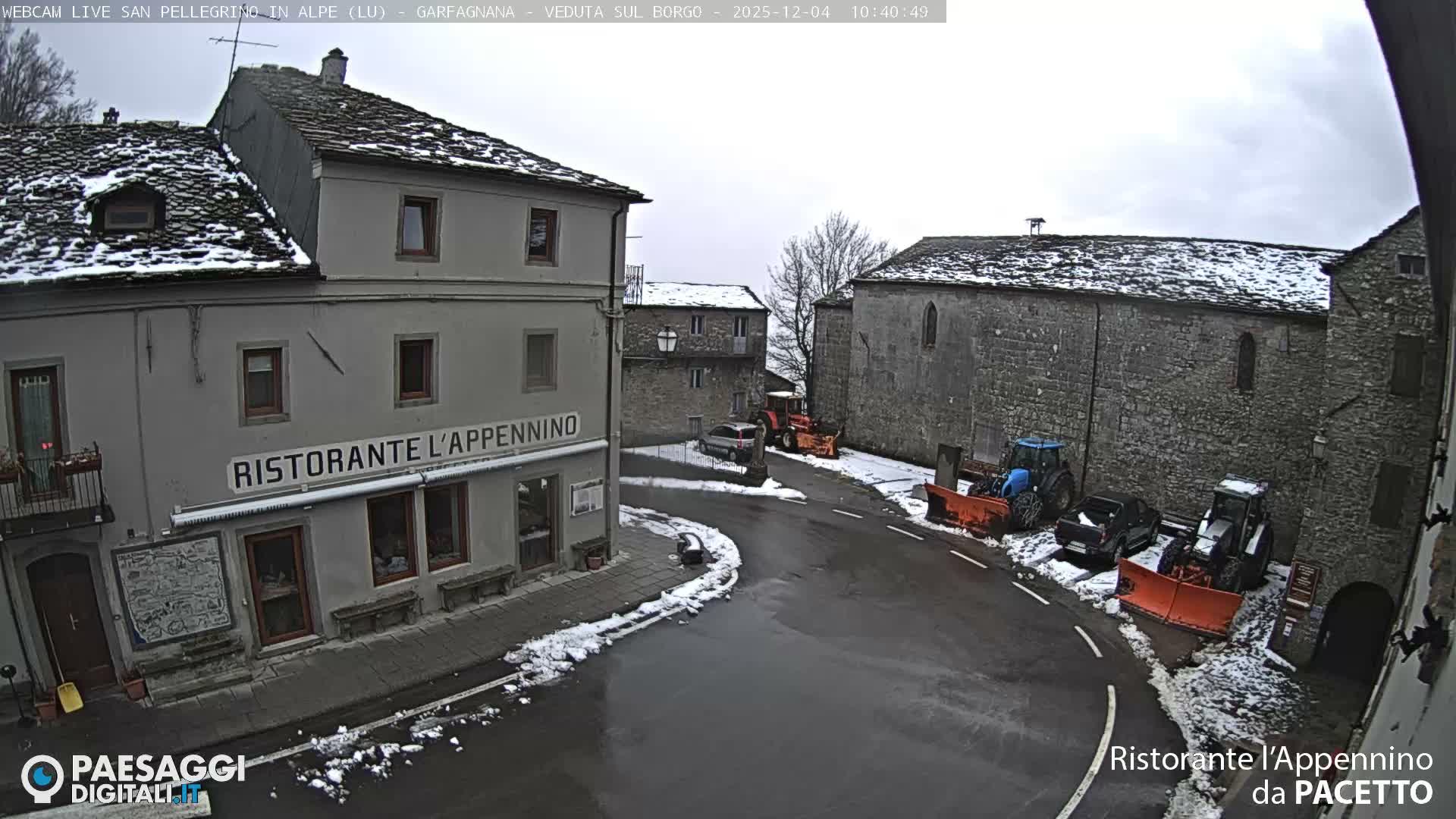 An overcast winter day reveals an old village scene with historic stone buildings, roofs lightly dusted with snow, a wet street, and multiple snowplows parked along the thoroughfare.