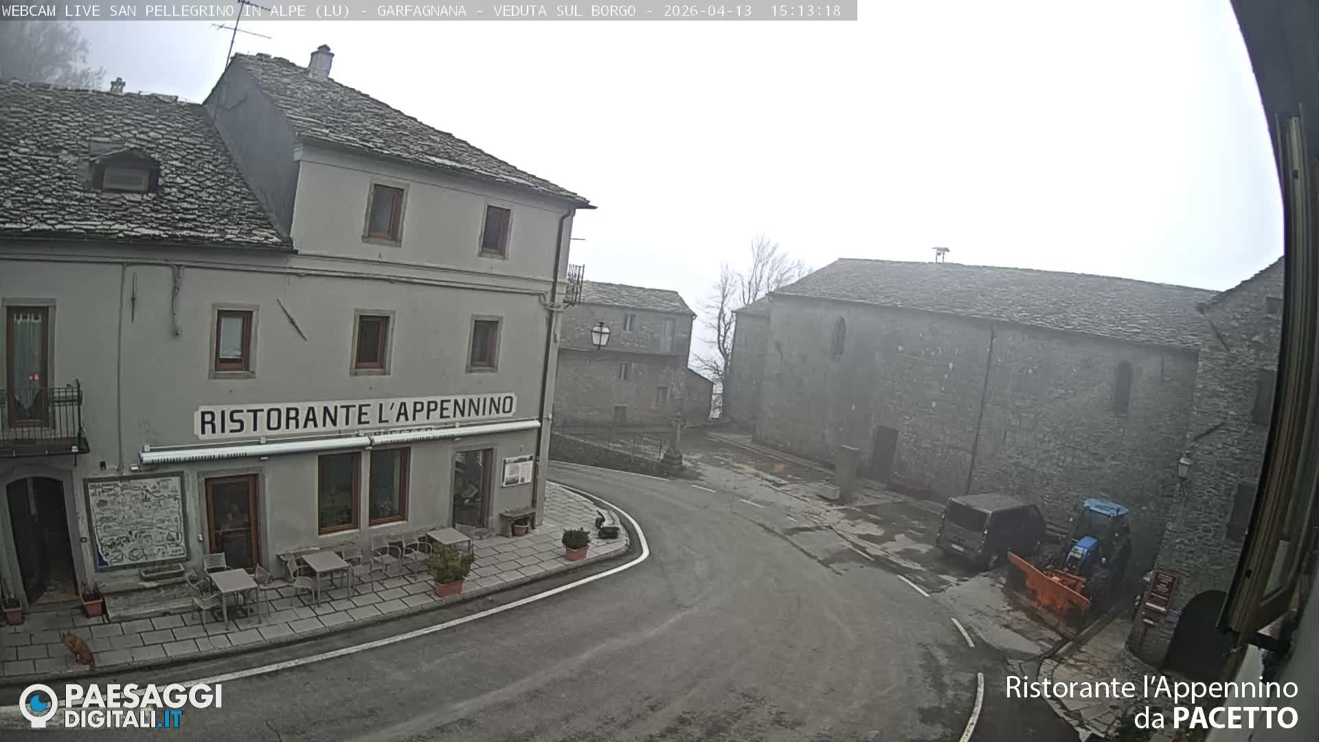 An overcast winter day reveals an old village scene with historic stone buildings, roofs lightly dusted with snow, a wet street, and multiple snowplows parked along the thoroughfare.