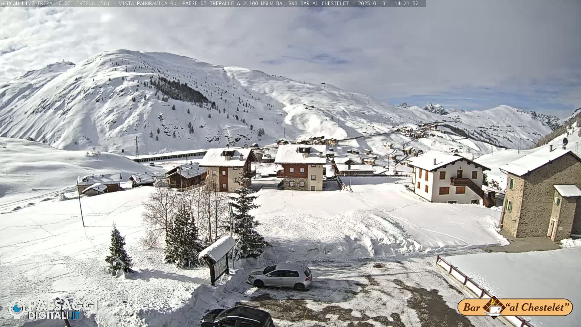 A snow-covered village nestled in a valley between snow-capped mountains on a partly sunny day.