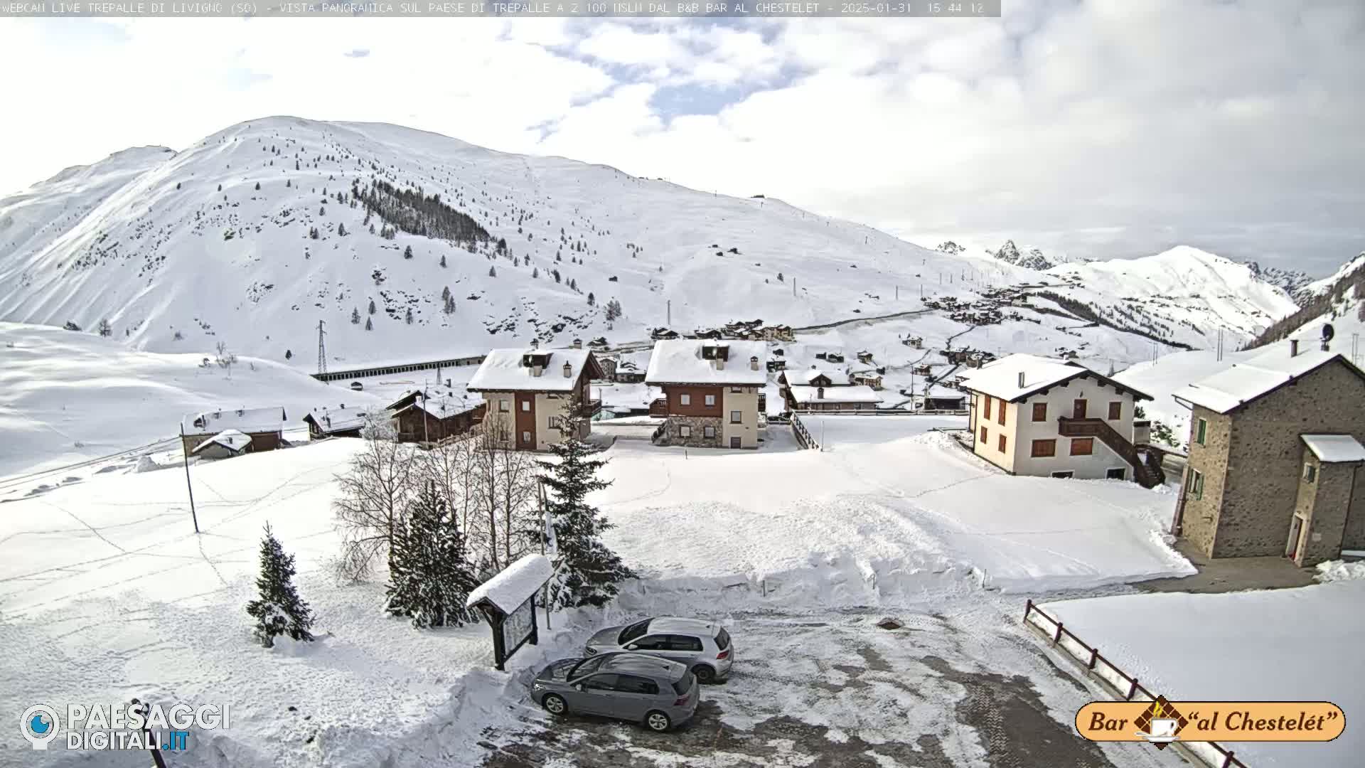 A snow-covered village nestled in a mountainous region under a partly cloudy sky, with two cars parked in a snowy lot.