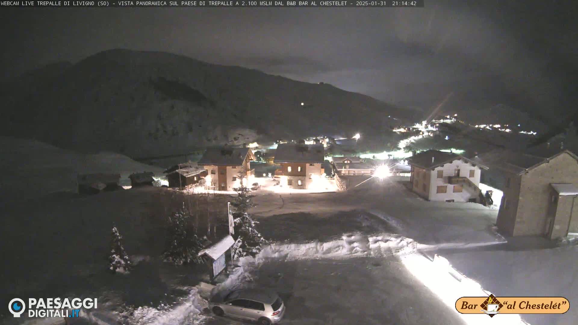A snow-covered village at night, illuminated by lights from buildings, is nestled in a valley surrounded by dark mountains under a cloudy sky.