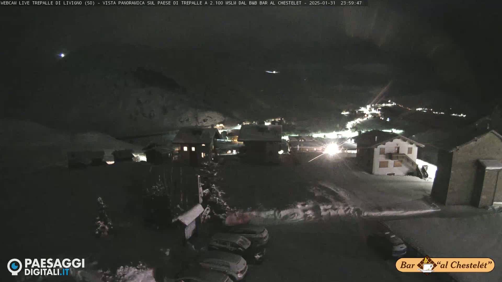 A nighttime, snow-covered village scene is illuminated by lights, with several cars parked in a lot, under a dark sky.