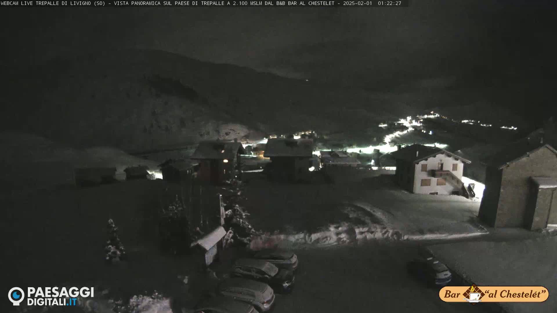 A nighttime, snow-covered village scene is illuminated by scattered lights, with cars parked in a lot in the foreground.