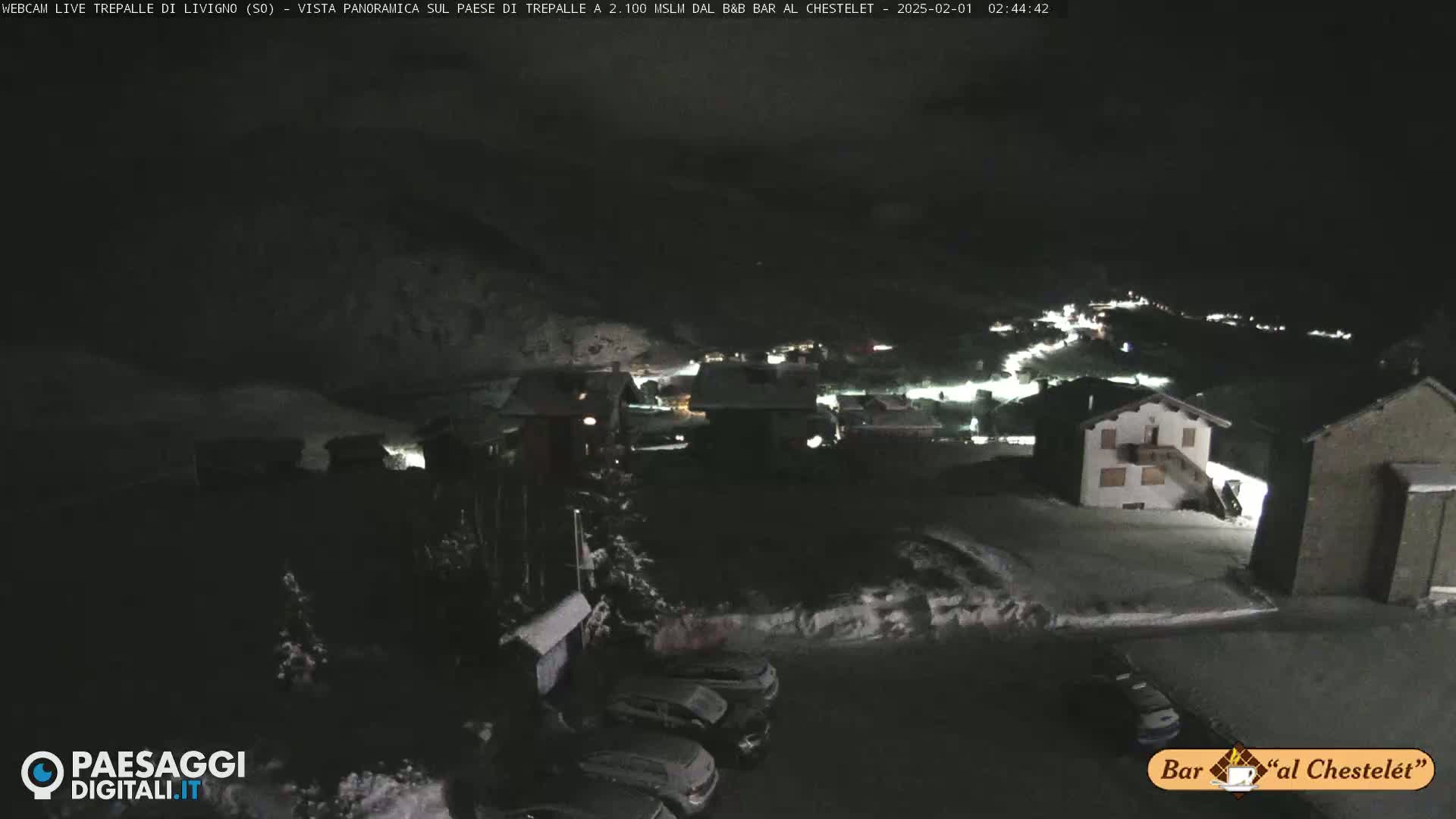 A nighttime, snow-covered village scene is illuminated by scattered lights, with several cars parked in a lot.
