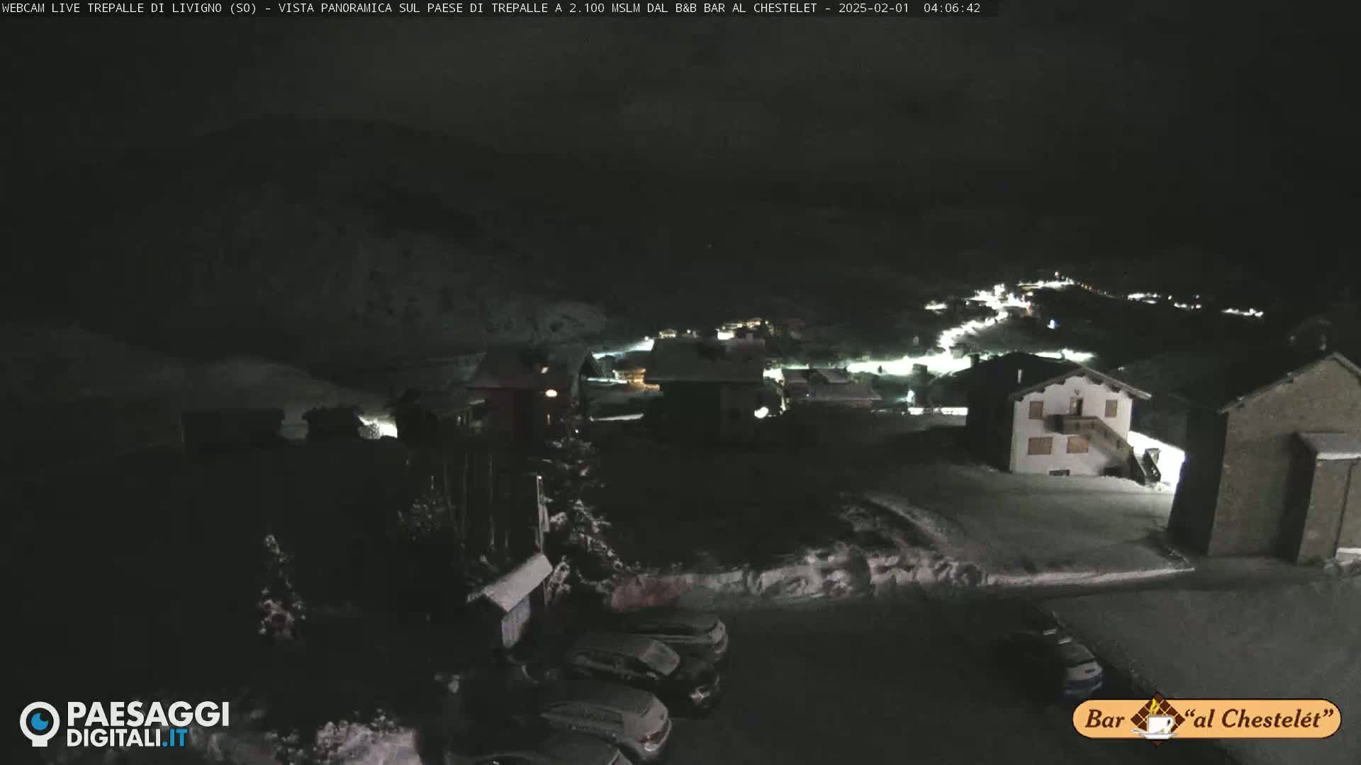 A nighttime, snow-covered village scene is visible, with several cars parked in a lot and some lights illuminating buildings.