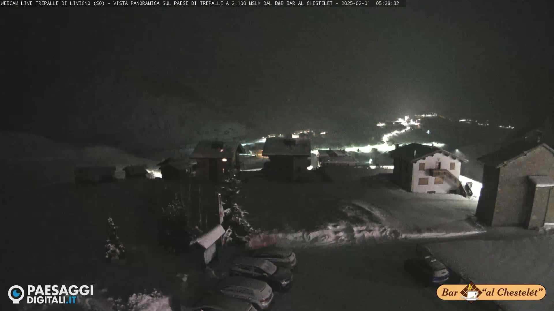 A snow-covered village at night is illuminated by scattered lights, with several cars parked in a lot.
