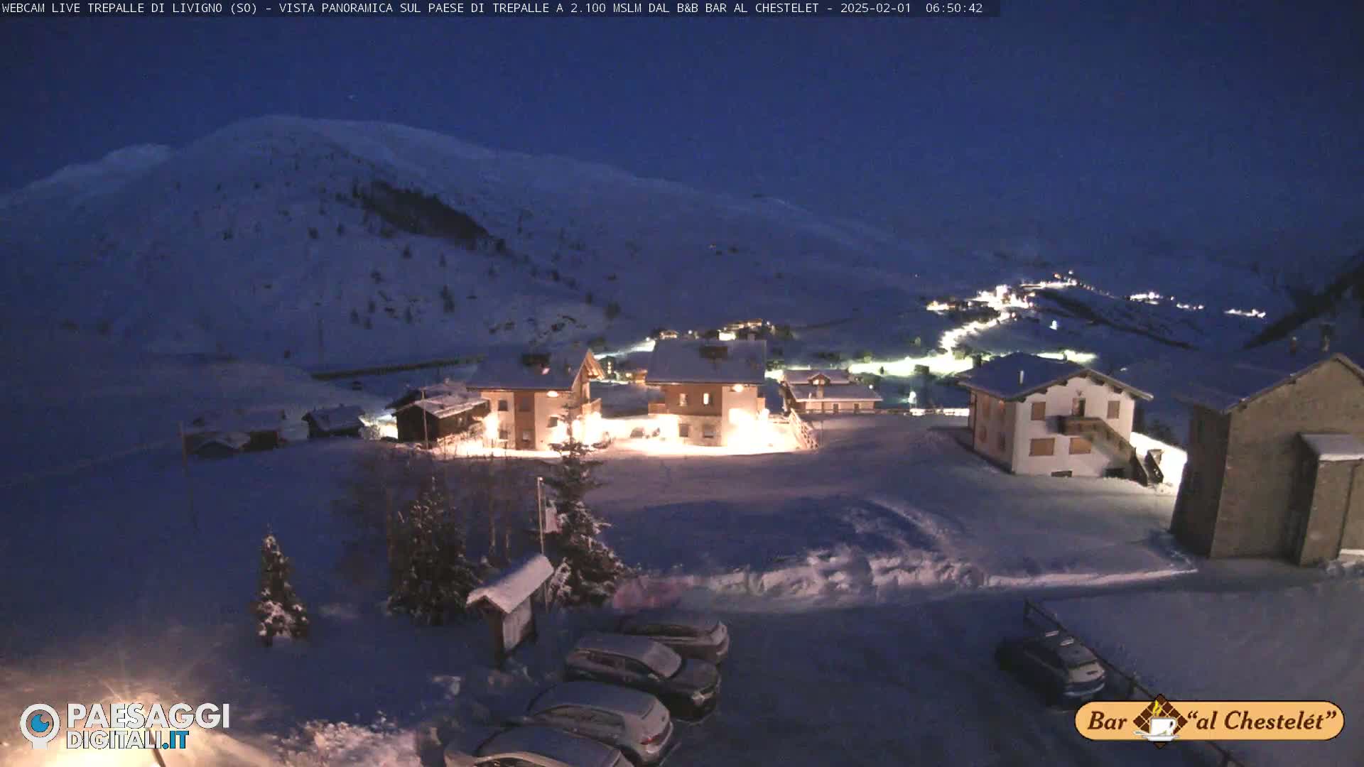 A snow-covered village is illuminated at night, nestled against a snow-covered mountain under a dark blue sky.