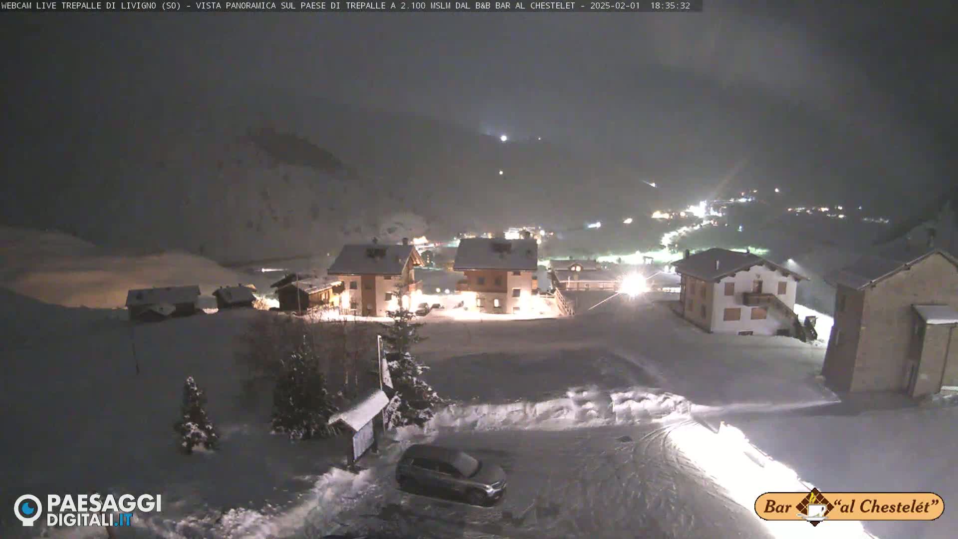 A snow-covered village is illuminated at night, with a car parked in a snowy lot in the foreground.