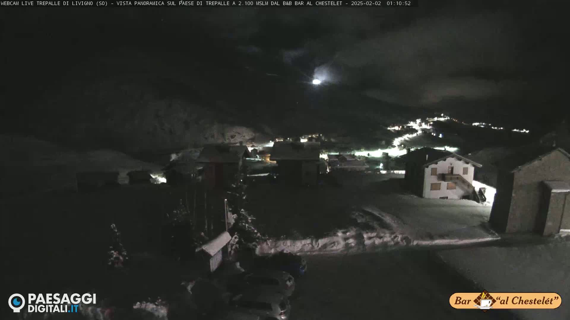 A nighttime, snow-covered village scene under a cloudy sky is illuminated by faint lights, with a few cars parked in a lot.