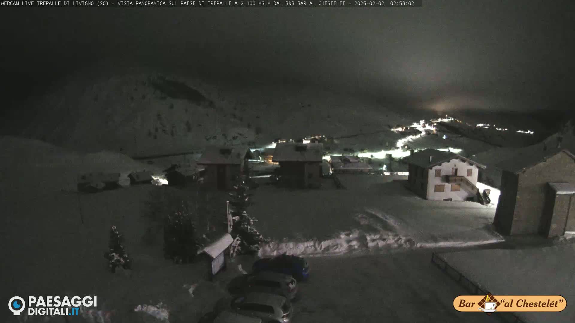 A snow-covered village at night, illuminated by scattered lights, is viewed from a slightly elevated position under a dark, cloudy sky.