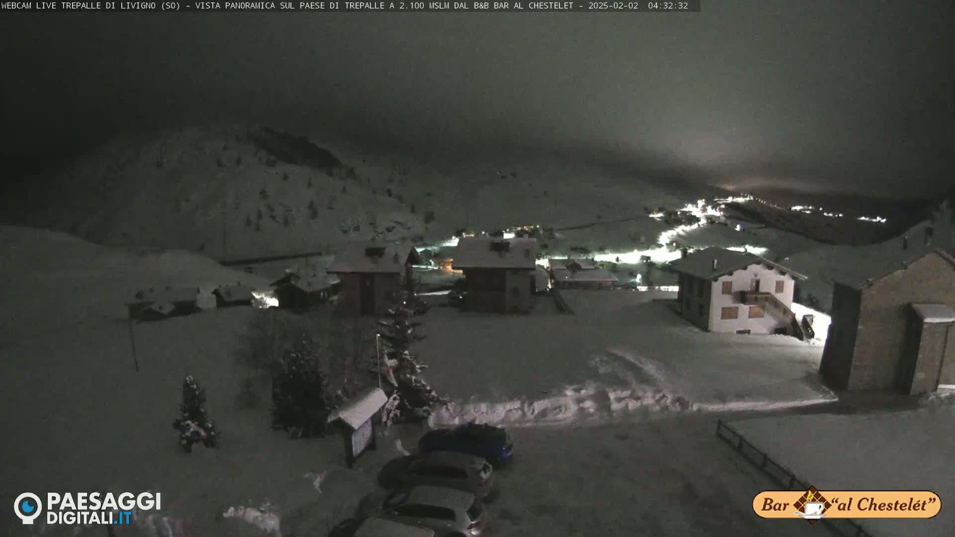 A snow-covered village at night is illuminated by scattered lights, with several cars parked nearby under a dark, overcast sky.