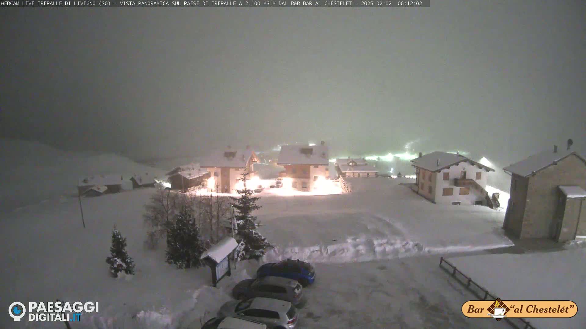 A snow-covered village at night is illuminated by lights from buildings, with several cars parked in a snowy lot, under a heavy snow.