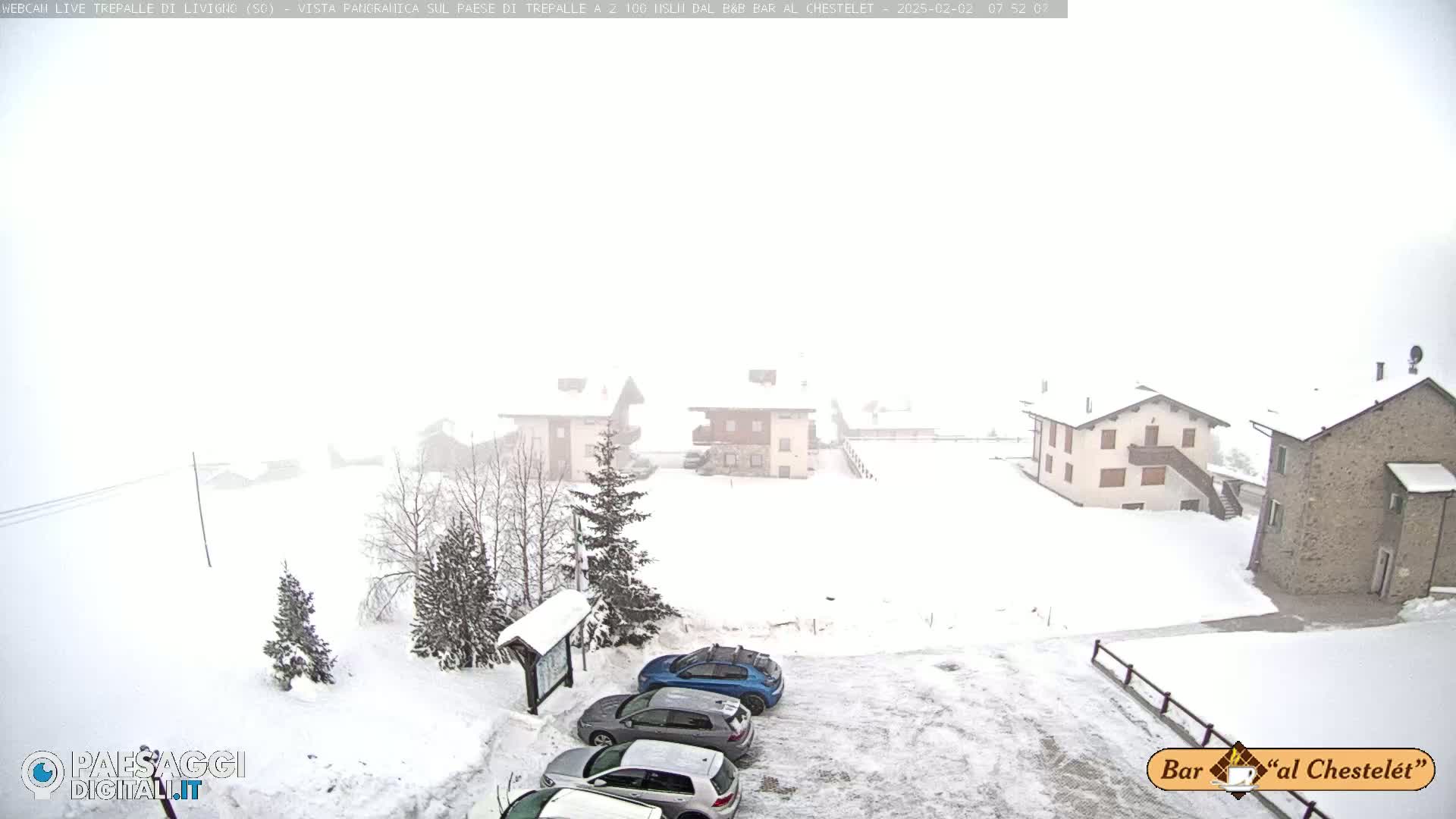A snow-covered parking lot with several cars and buildings in the background during a heavy snowstorm.
