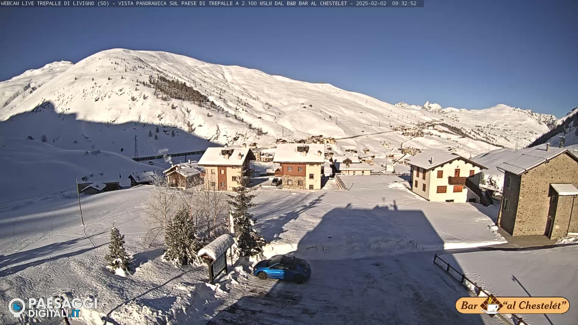 A snow-covered village nestled in a valley beneath a sunny, snow-capped mountain.