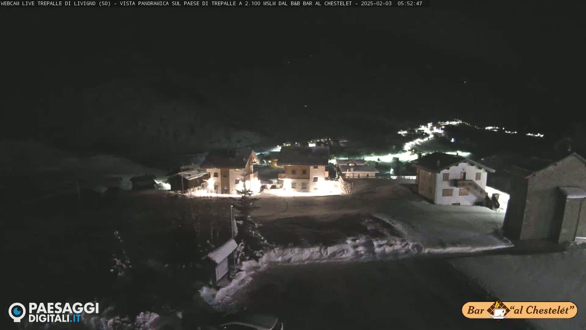 A nighttime, snow-covered village is illuminated by building lights under a dark sky.