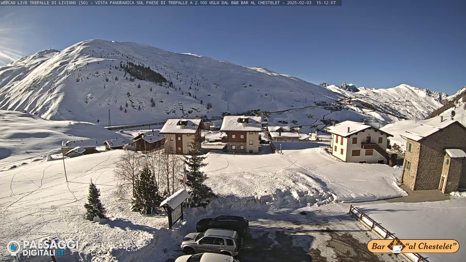 A snow-covered mountain village scene under a clear, sunny sky, with several buildings and cars visible in a parking lot.