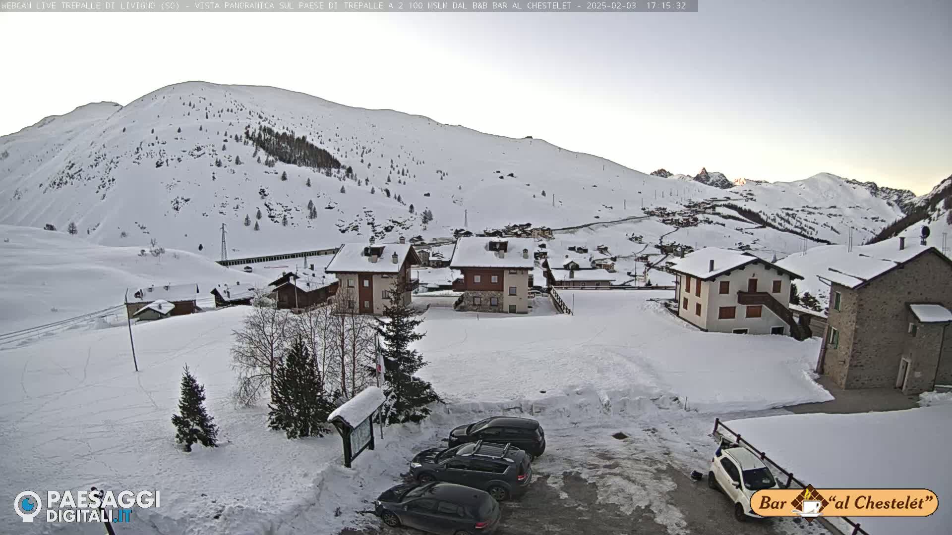 A snow-covered mountain village with several buildings and a few cars parked in a snowy lot, under a clear, bright sky.