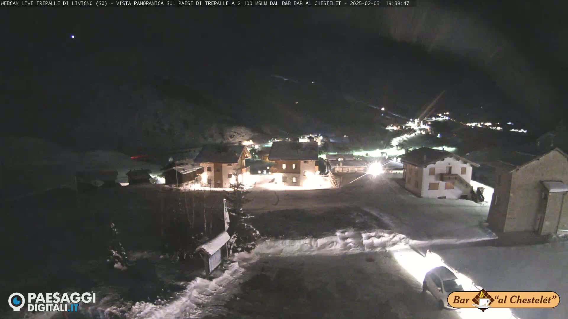 A nighttime, snow-covered village is illuminated, with several buildings visible under a clear, dark sky.