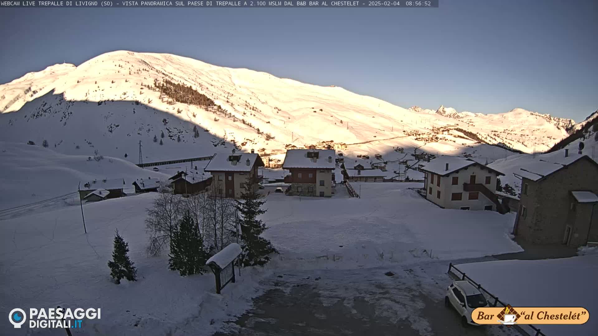A snow-covered village nestled at the base of a sunlit, snow-dusted mountain under a clear blue sky.