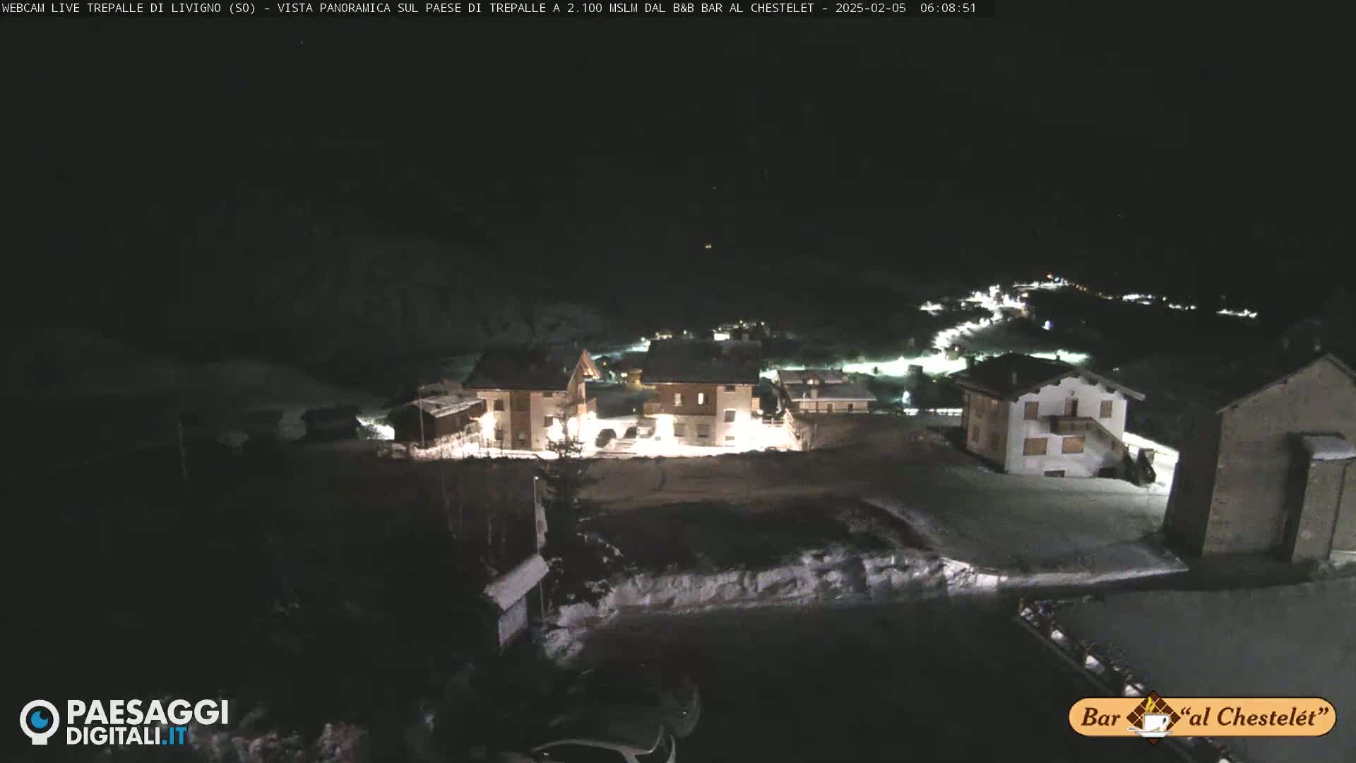 A nighttime, snow-covered village is illuminated by the lights of its buildings under a dark sky.