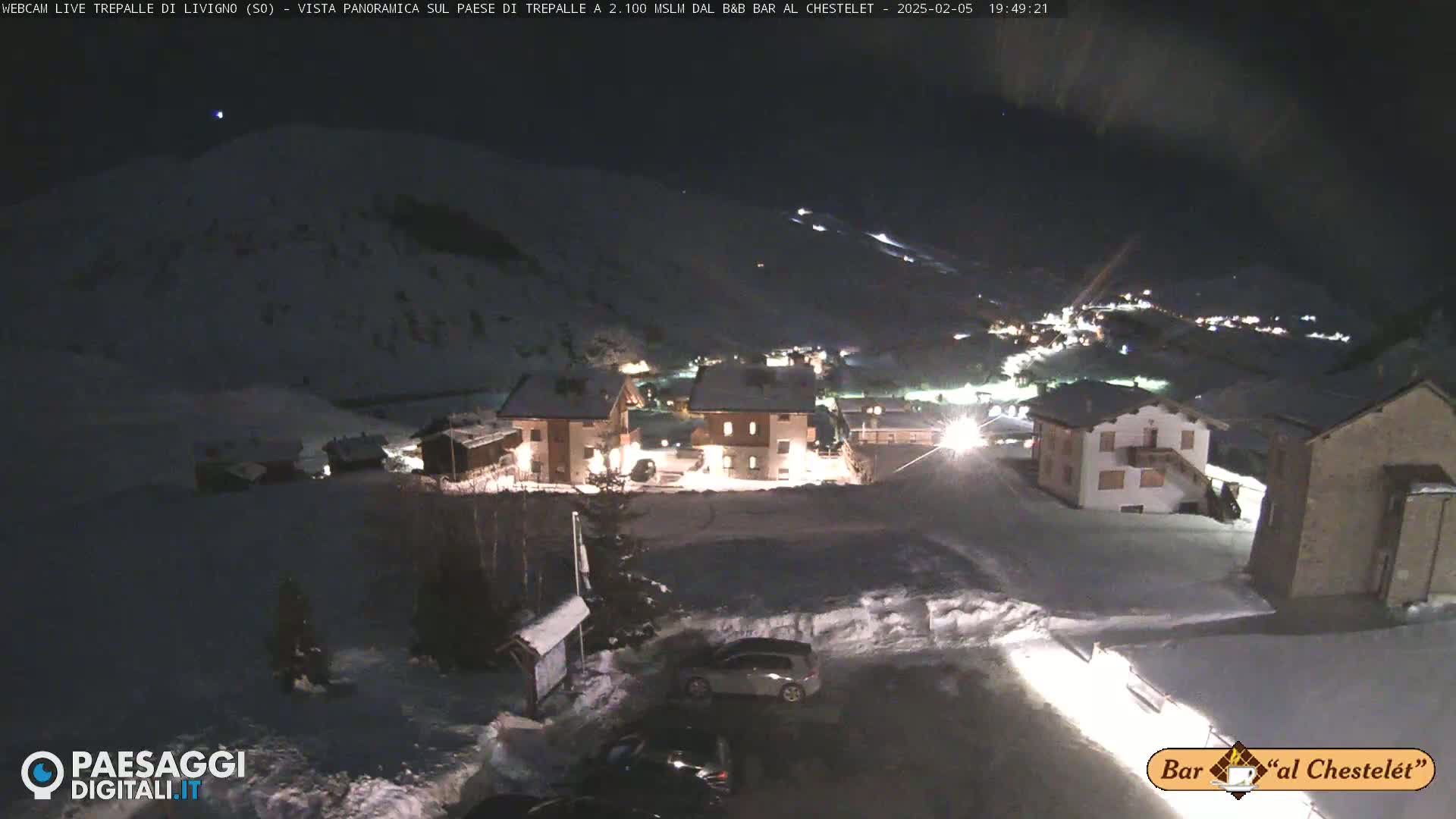 A snow-covered village is illuminated at night, with a few cars parked in a snowy lot.