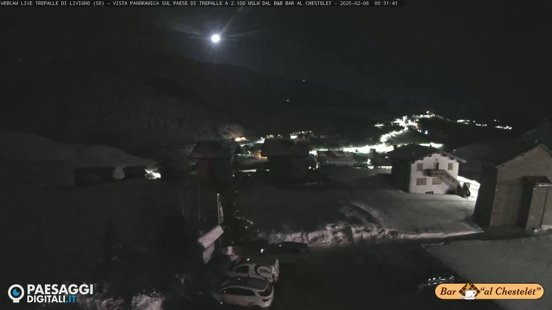 A nighttime, snow-covered village is illuminated by a bright moon and scattered lights, with a few vehicles parked near buildings.