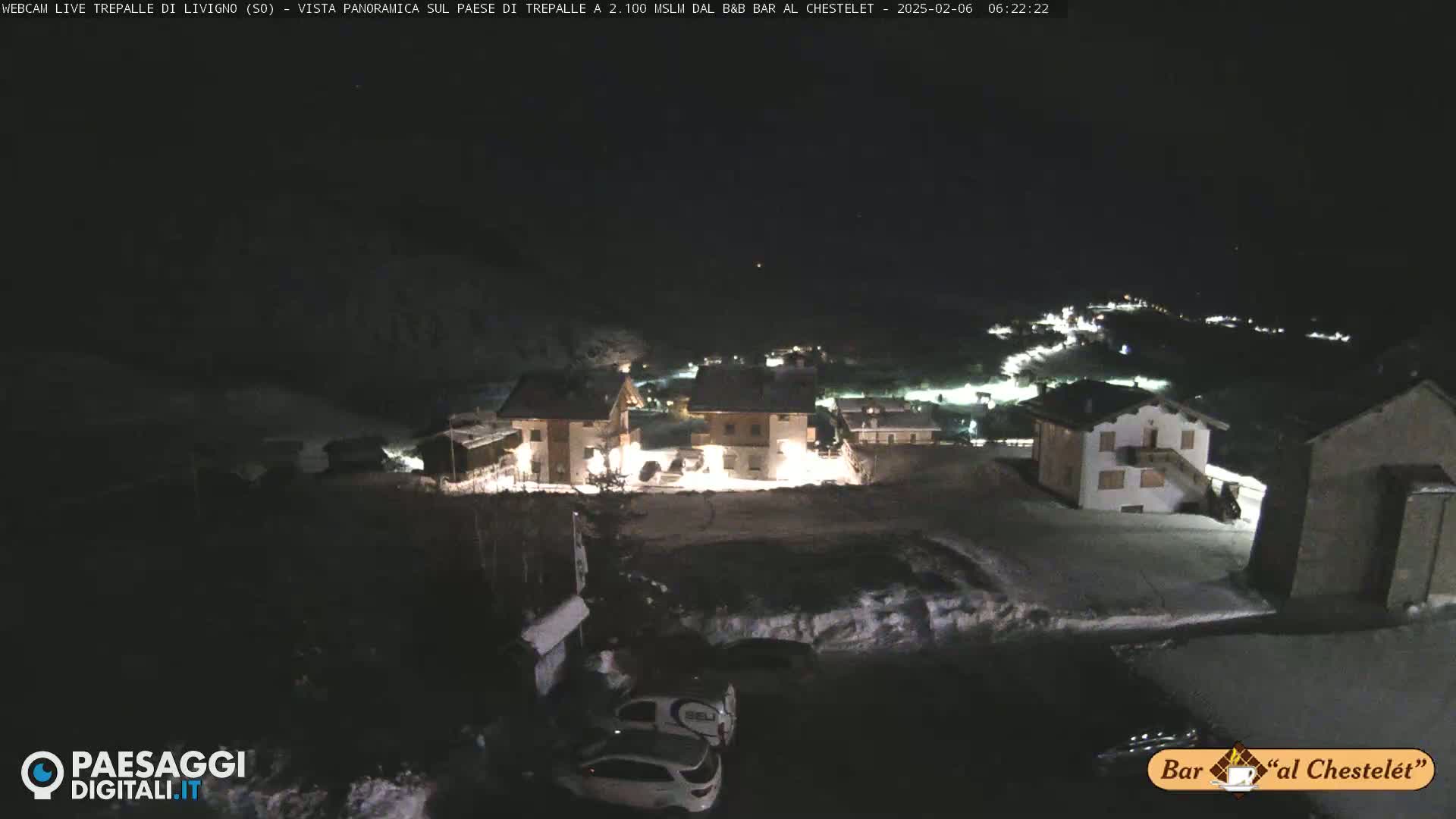 A nighttime, snow-covered village scene with several illuminated buildings, a few parked cars, and a dark, clear sky.