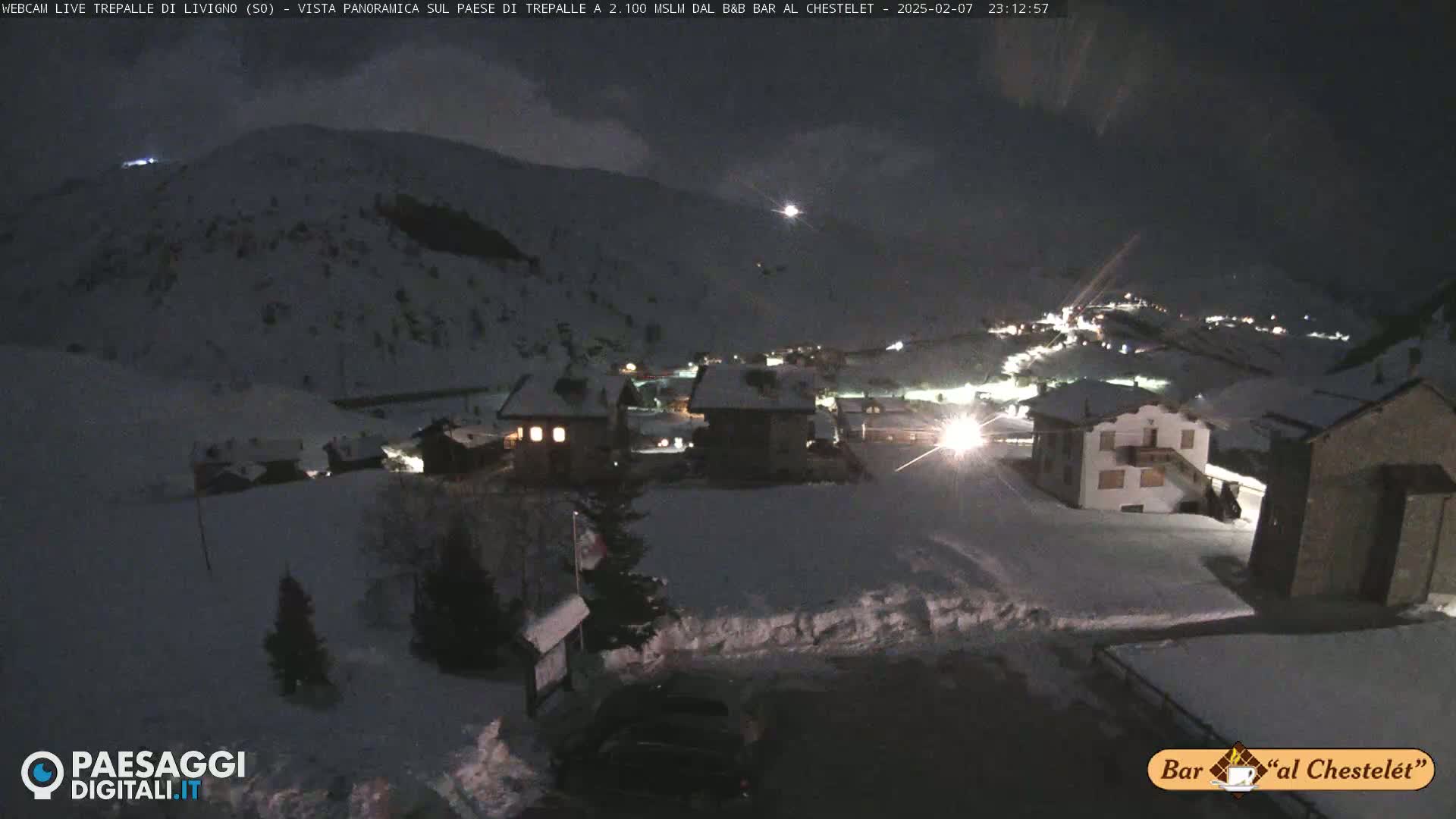 A snow-covered village at night, illuminated by lights from buildings nestled against a dark, snow-dusted mountainside under a mostly clear night sky.