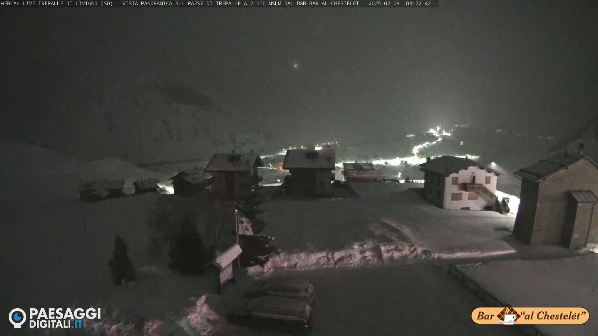 A snow-covered village at night, illuminated by scattered lights, is seen under a dark, possibly cloudy sky.