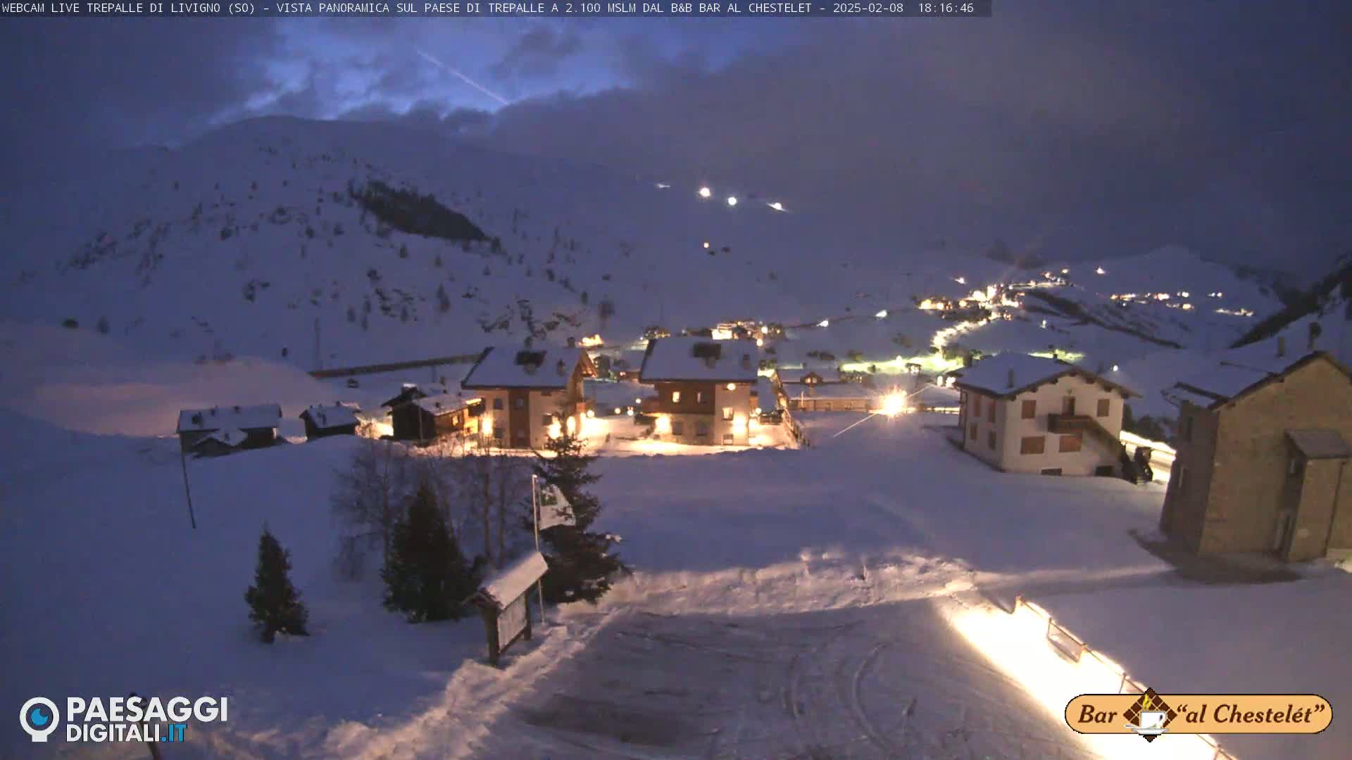 A snow-covered village at night, illuminated by lights from buildings, sits on a hillside under a dark, twilight sky.